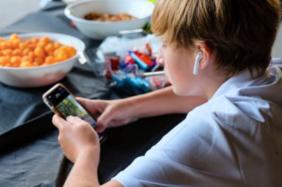 Preteen boy concentrating on smartphone with ear buds and bowls of snacks