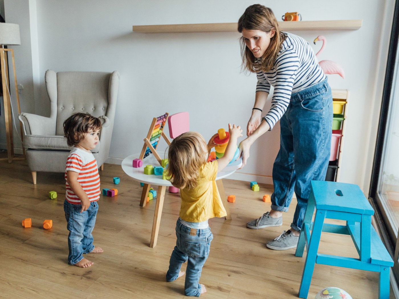 Positive young female babysitter in casual clothes playing with infant children in cozy playroom with colorful educational toys
