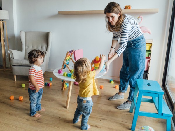 Positive young female babysitter in casual clothes playing with infant children in cozy playroom with colorful educational toys
