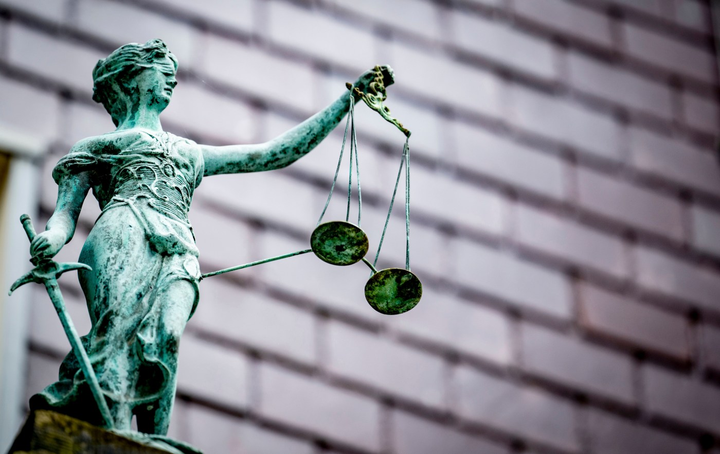 A weathered statue of Lady Justice holding the scales of justice against a blurred building backdrop