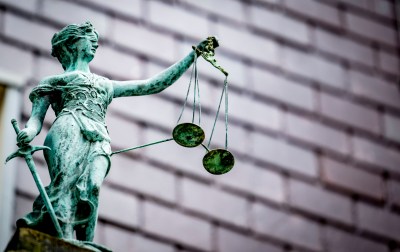 A weathered statue of Lady Justice holding the scales of justice against a blurred building backdrop